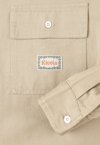 Camicia beige intrecciata con polsino abbottonato. La tasca sul petto sinistro presenta un'etichetta con la scritta "Rivvia" in arancione, bordata da un motivo floreale.