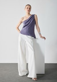 Asimetriška violetinė palaidinė iš lino, derinama su baltomis plačiomis kelnėmis; modelis dėvi sidabrinį apyrankę ir stovi šalia baltos kolonos.