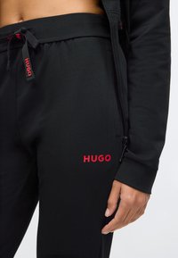 Black jogger pants made from soft fabric, featuring a red "HUGO" logo on the left thigh and a drawstring waist for adjustable fit.