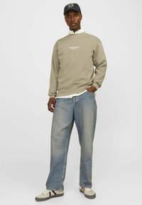Sudadera verde oliva con logo blanco, jeans azules de corte holgado, zapatillas grises y una gorra negra con diseño bajo y acentos mínimos.