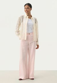 Spitzenhemd in Creme mit Diamantmuster, weißes Tanktop und hellrosafarbene, weit geschnittene Leinenhose. Braune Schuhe runden das Outfit ab.