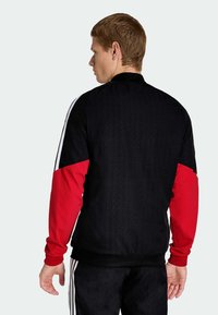 Veste de survêtement noire et rouge avec des panneaux unis, des rayures blanches sur les manches, un tissu texturé et une coupe ajustée.