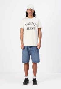 Šviesiai kreminės spalvos marškiniai su "TOMMY JEANS" logo, džinsiniai šortai ir juodi sportbačiai. Modelis taip pat dėvi baltą kepurę, stovi prieš baltą foną.