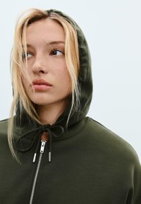 Young person with light blonde hair wearing an olive green zip-up hoodie with the hood up, looking to the side against a plain background.