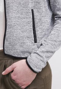 Personne portant une veste grise texturée avec des bordures noires, la main placée dans la poche d'un pantalon vert olive.