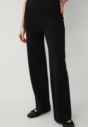 Kvinde iført sorte bukser med vide ben og sorte balletsko, stående med den ene hånd på hoften mod en lys baggrund.