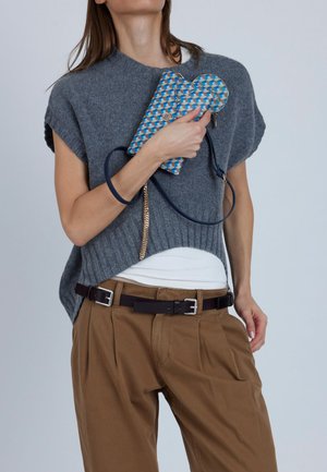 Sivý pletený top, hnedé plisované nohavice a modrý listový vak s geometrickým vzorom a retiazkou na rameno. Viditeľné detaily vrstveného, neformálneho outfitu.