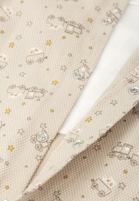 Tela beige con un tejido texturizado, que presenta un estampado de trenes y estrellas en marrón y dorado. Botones a presión plateados detallan el cierre.