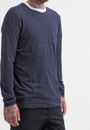 Man draagt een marineblauw gespetterd shirt met lange mouwen, een klein borstzakje en zwarte broek, met zichtbare getatoeëerde handen en nek.