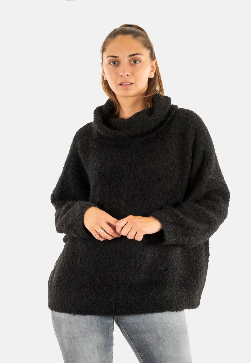 Junge Frau mit zurückgebundenem Haar, die einen schwarzen strukturierten Rollkragenpullover und hellgraue Jeans trägt und mit locker gefalteten Händen dasteht.
