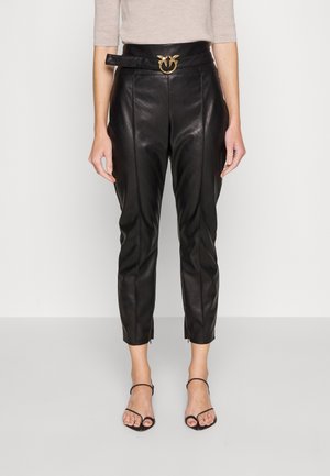 Kobieta w czarnych, sztucznych skórzanych spodniach z wysokim stanem, z paskiem z złotą klamrą, beżowej bluzce oraz czarnych, odkrytych butach na obcasie z paskami, stojąca na tle białego tła.