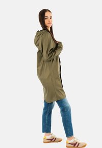 Chaqueta de lluvia ligera en verde oliva con capucha, cierre frontal con cremallera, bolsillos laterales, combinada con unos jeans azules y zapatillas beige con detalles en burdeos.