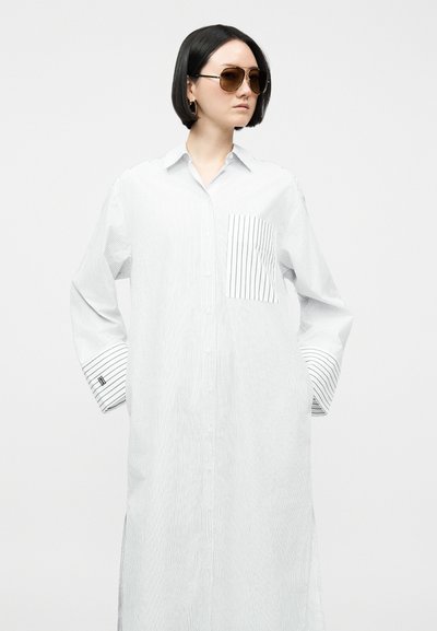 Γυναίκα με κοντά μαύρα μαλλιά που φοράει γυαλιά aviator ηλίου και ένα μακρύ λευκό πουκάμισο-φόρεμα με λεπτές μαύρες κάθετες ρίγες και τσέπη στο στήθος με ρίγες.