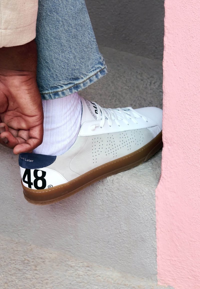 Personne portant une basket blanche avec le numéro 48 au talon, une chaussette blanche et un jean bleu, ajustant sa chaussure tout en posant le pied sur une marche en béton gris.