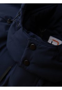 Detailní záběr na límec námořnické modré bundy se dvěma černými patentními knoflíky a nastavitelným šňůrkovým poutkem.