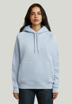 Frau mit langen dunklen Haaren und Sommersprossen, die eine hellblaue Kapuzenjacke und dunkle Jeans trägt, stehend vor einem einfarbigen hellen Hintergrund.