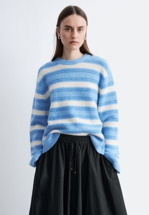 Jonge vrouw met stijlbruin haar draagt een blauw-wit gestreepte trui en een zwarte rok, staand tegen een lichte achtergrond.