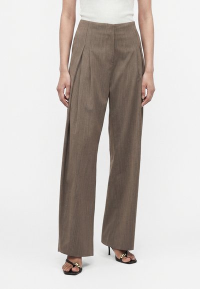 Femme portant un pantalon marron taille haute à jambes larges et des talons ouverts noirs avec des accents dorés, debout devant un fond blanc uni.