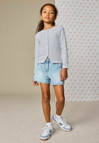 Camiseta de manga larga a rayas con cierre de botones, pantalones cortos de mezclilla azul claro y zapatillas grises con detalles blancos. Fondo con patrón floral.