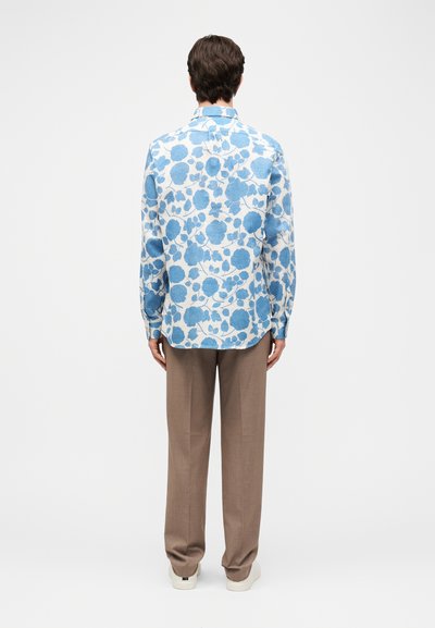 Άνδρας που στέκεται γυρισμένος πλάτη, φορώντας λευκό πουκάμισο με μπλε λουλουδάτο μοτίβο, καφέ παντελόνι και λευκά αθλητικά παπούτσια σε απλό φόντο.