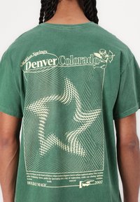 Groene katoenen t-shirt met een relaxte pasvorm, voorzien van crème-kleurige tekst en een zwervende stergrafiek op de achterkant, samen met een vintage-stijl illustratie.