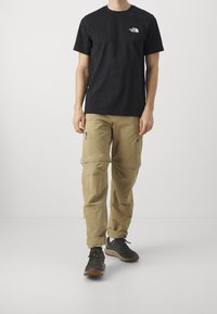 Czarny bawełniany t-shirt z logo na piersi, zestawiony z beżowymi spodniami typu cargo z kieszeniami na zamek oraz szarymi butami trekkingowymi z teksturowaną podeszwą.