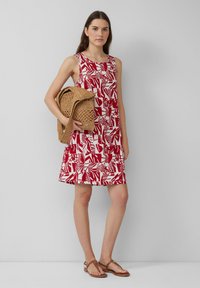 Czerwona i biała sukienka bez rękawów z abstrakcyjnym wzorem liści, w kształcie rozszerzanym, zestawiona z plecioną brązową torbą i brązowymi sandałami.