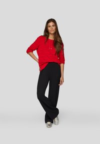 Frau steht mit der Hand auf der Hüfte, trägt ein strukturiertes rotes Langarmoberteil, lockere schwarze Hose und silberne Sneakers vor einem grauen Hintergrund.