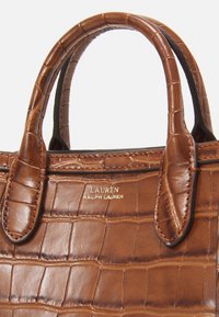Brązowa torebka skórzana z tłoczeniem w kształcie krokodyla z podwójnymi uchwytami, z złotą tabliczką logo. Gładka tekstura i usztywniony kształt.