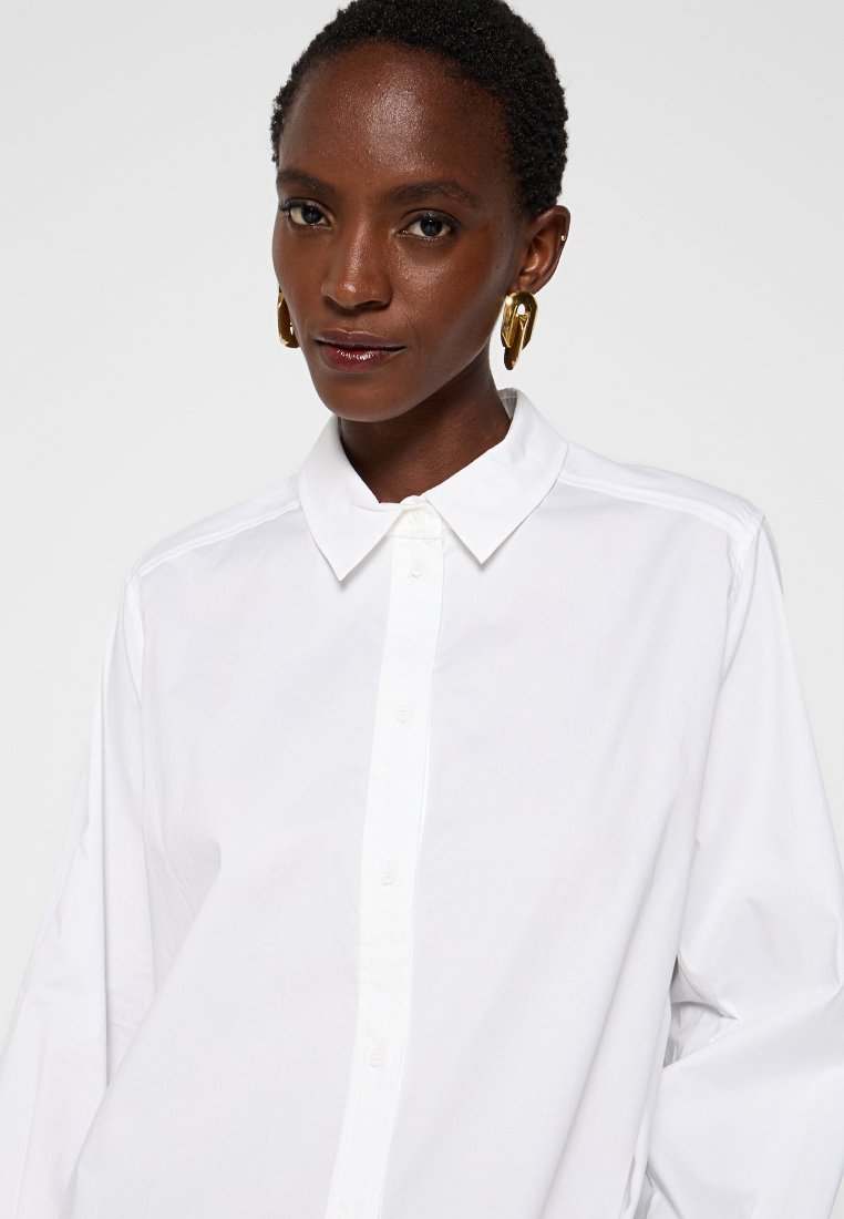 Witte button-up shirt met een kraag, gemaakt van gladde stof. Heeft een volledige knoopsluiting en lange mouwen, gedragen met gouden oorbellen.