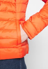 Lyseorange polstret jakke med vandrette syninger, med en sidelomme og tætsiddende ærmer. Materialet virker glat og let.