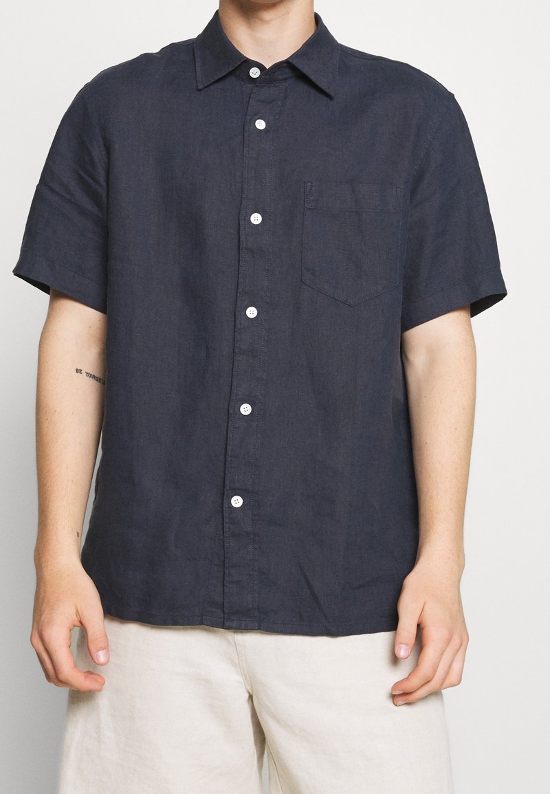 Homme portant une chemise bleu marine foncé à manches courtes avec une poche poitrine et un pantalon beige clair, debout devant un fond uni.