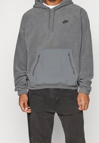 Šedý fleecový pulovr s kapucí na šňůrku, přední kapsy na zip a elastickým lemem. Na hrudi je černé logo Nike.