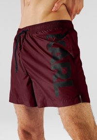 Bañador burdeos con un estampado de logo negro. Hecho de tela ligera con cintura elástica y cordón. Incluye aberturas laterales.