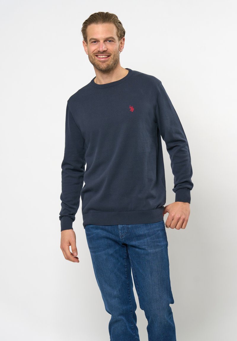 Marineblå sweater i ribstrik, med rund hals og et lille rødt logo på brystet. Parret med denim jeans.