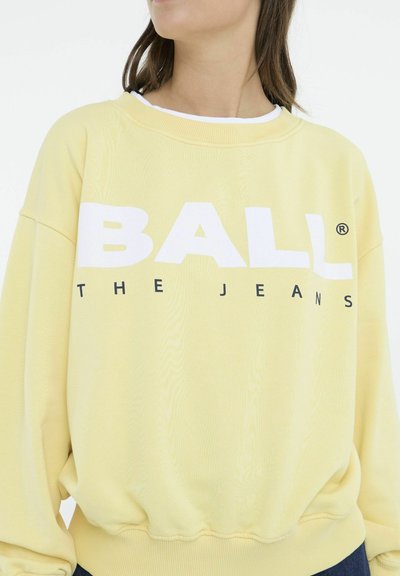 Gul sweatshirt med løs pasform, der har fed hvid tekst "BALL" og "THE JEANS" trykt på forsiden. Blødt, struktureret stof.