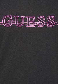 Černé tričko s velkým růžovým logem "GUESS" a vodorovnou čárou přes text. Tkanina vypadá hladce a elasticky.