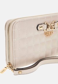 Beige gesteppte Guess Geldbörse mit goldenen Reißverschluss, goldener Logo-Plakette und angebrachtem Handgelenkband mit goldenen Nieten.