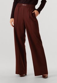 Pantalons larges bordeaux avec une taille élastique et des poches latérales. Tissu lisse avec un tombé subtil et des plis à l'avant pour une forme supplémentaire.