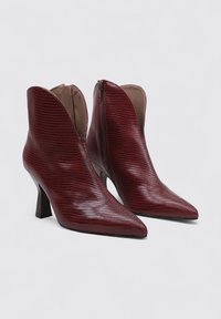 Botines de tobillo en color marrón, texturizados, con punta afilada y tacón de aguja alto. Presentan un cierre lateral con cremallera y un diseño distintivo de recortes curvados.