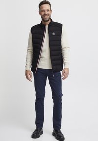 Svart puffervest med glidelås, røde og hvite aksentstriper, beige strikkegenser under, marineblå bukser og svarte sko.