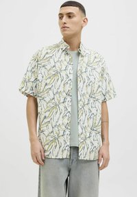 Kortærmet skjorte i let stof med et gult og blåt bladmønster på en cremefarvet baggrund, iført over en lysegrøn T-shirt.