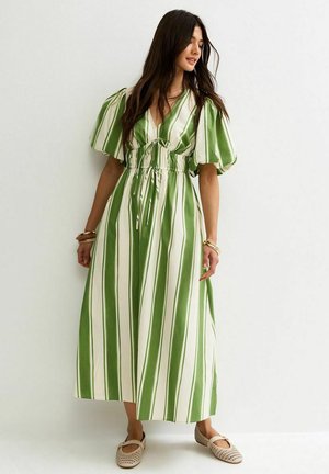 Kobieta w sukience w pionowe zielono-białe paski z bufkami na rękawach, taliowana, w beżowych płaskich butach, stojąca na tle białej ściany.