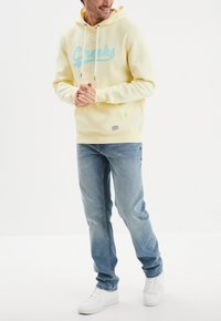 Felpa gialla con logo blu "Creeks", tasca frontale, cappuccio con cordino, abbinata a jeans azzurri chiari e sneakers bianche. Textura liscia.
