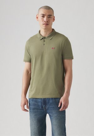 Oliwkowa koszulka polo wykonana z bawełny, z trzema guzikami na przodzie i małym czerwonym logo na lewej piersi. Nosi ją z niebieskimi dżinsami.