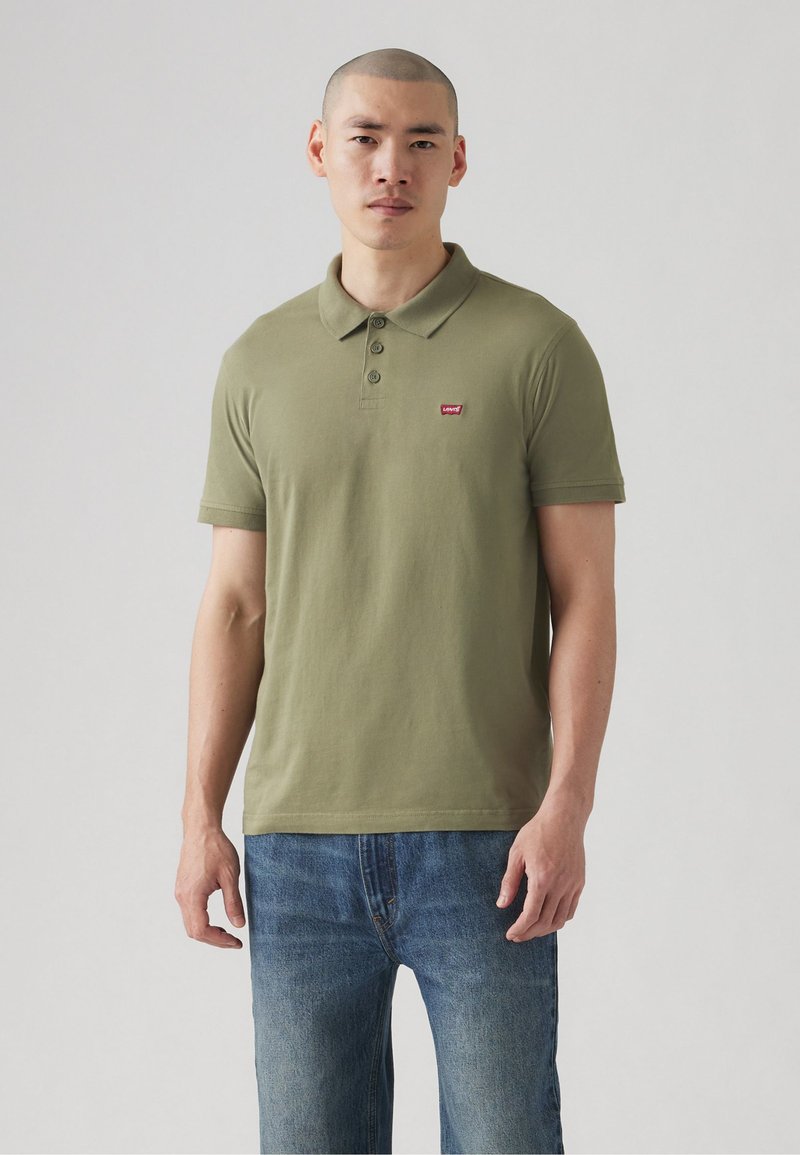 Polo de culoare verde măslin, fabricat din bumbac, având o deschidere cu trei butoane și un mic logo roșu pe pieptul stâng. Purtați cu blugi albaștri.