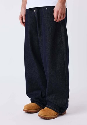 Homme portant un pantalon en jean bleu foncé oversize et des bottes en daim beige, debout sur un fond blanc.