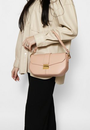 Donna con camicia beige oversize e gonna nera tiene una borsa in pelle rosa chiaro con chiusura dorata sul braccio.