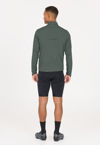 Vindjakke i mørkegrøn, letvægtsmateriale. Sammen med sorte polstrede cykelshorts og sorte sokker; skoene har en struktureret overflade.