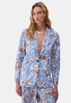 Donna con lunghi capelli biondi che indossa un completo blu a fiori marroni e bianchi, e una collana d'oro su sfondo grigio.